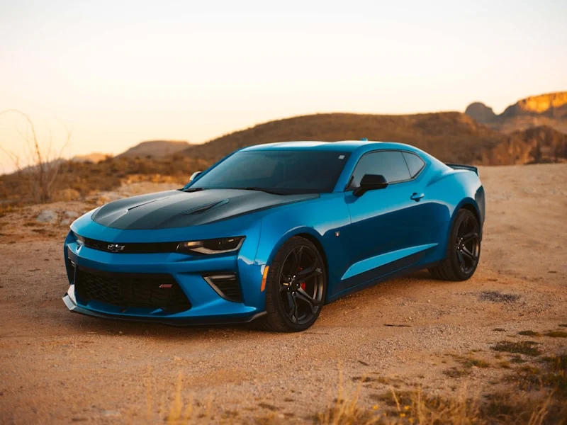 Een blauwe Chevrolet Camaro geparkeerd in een woestijnlandschap bij zonsondergang.