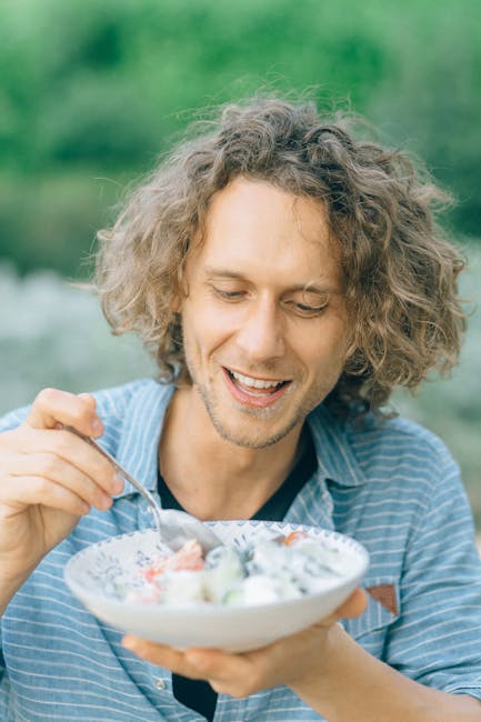 Smiling man with curly hair enjoys a bowl of fresh salad outdoors. Bright and lively summer scene.