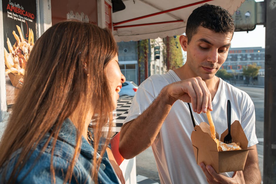 Two young adults savoring fries from a food truck outdoors on a sunny day.