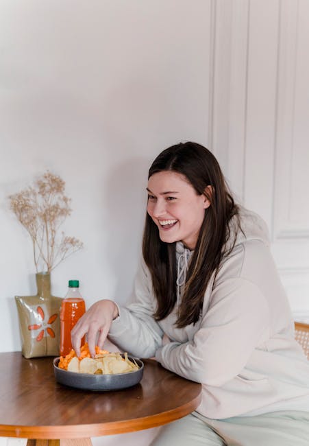 Positive woman with long brown hair in casual clothes sitting at wooden table with soda and eating tasty chips at home in daytime
