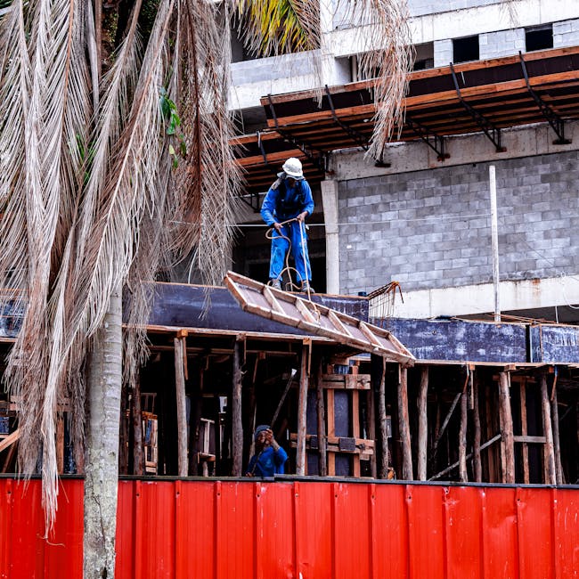 Bouwvakkers werken aan een constructie op een bouwplaats met een rode wand op de voorgrond.