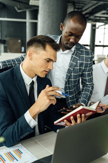 Two businessmen discussing work in a modern office setting with a laptop and documents.