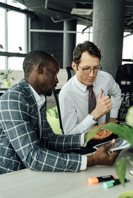 Two businessmen discussing work at a desk in a contemporary office setting.