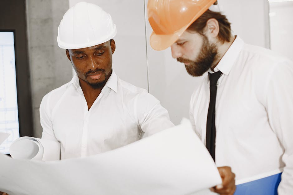 Two engineers in hard hats concentrate on reviewing detailed construction blueprints indoors.