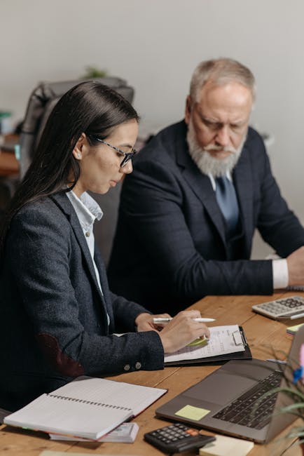 Two professionals in an office setting analyzing documents and using laptops.