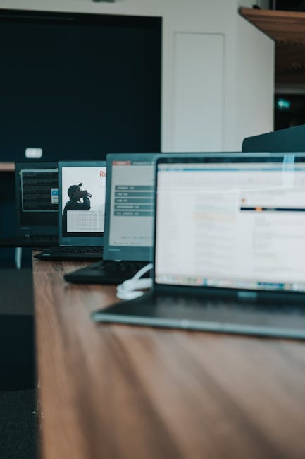 A line of laptops with screens on a wooden table in a modern office setting.