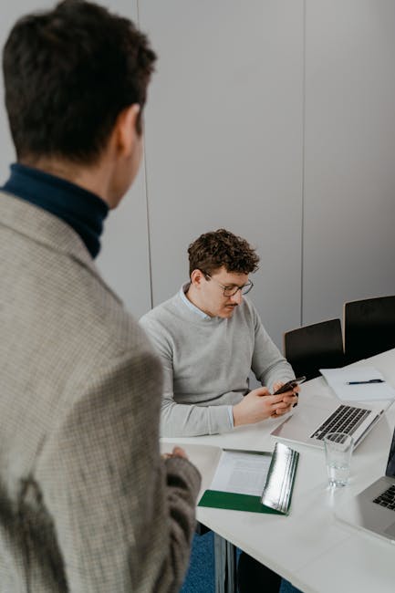 Two colleagues engage in a business meeting using laptops and smartphones in a modern office setting.
