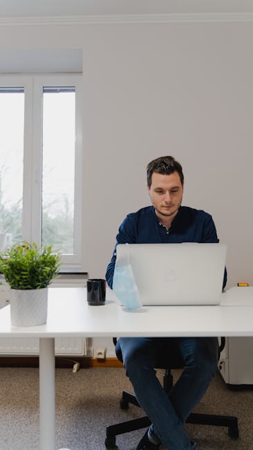 Adult man focused on laptop work at home office desk with coffee and plant.
