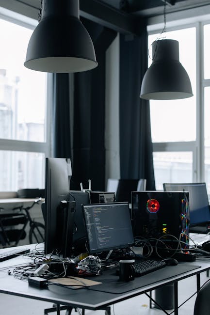 A modern office desk with computers, monitors, and tech equipment in a bright room.