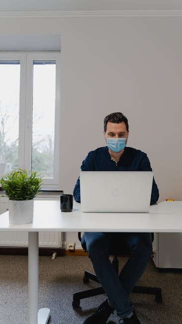 Man wearing a face mask working on a laptop from home with a potted plant on the desk.