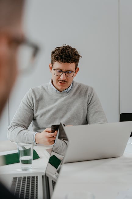Young man in glasses and gray sweater focused on smartphone at work desk with laptop.