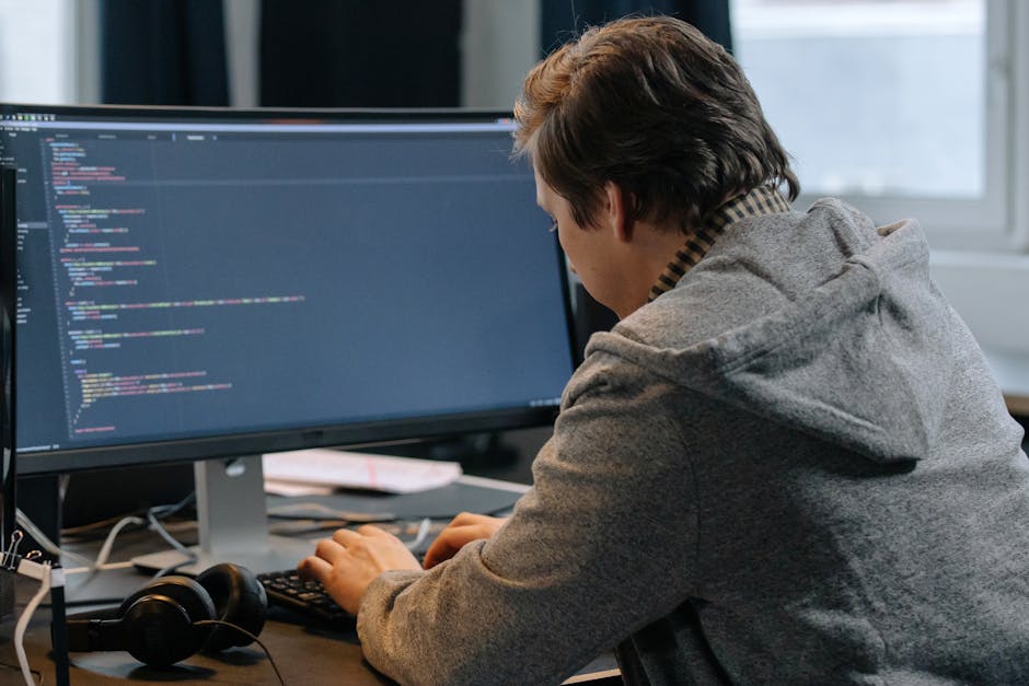 A man in a hoodie coding at a desk with large monitors in an office setting.