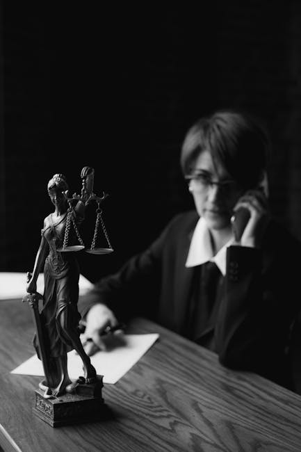A black and white photo of a lawyer on the phone with a Lady Justice figurine in focus.