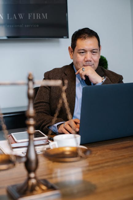 Cheerful ethnic legislator in formal outfit working with netbook at table with scales of justice and tablet