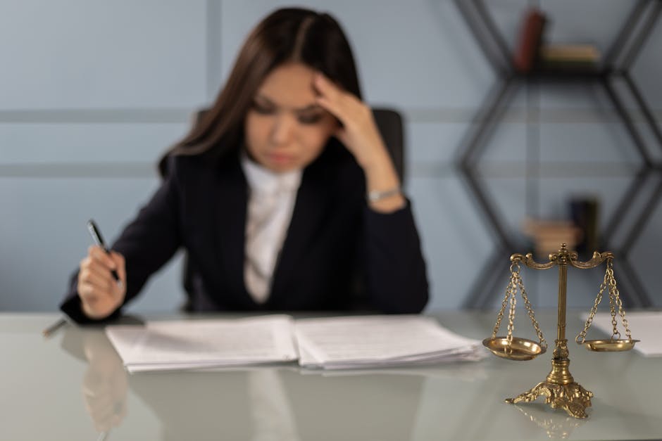 A fatigued female lawyer reviewing documents with a justice scale nearby, emphasizing stress in legal work.