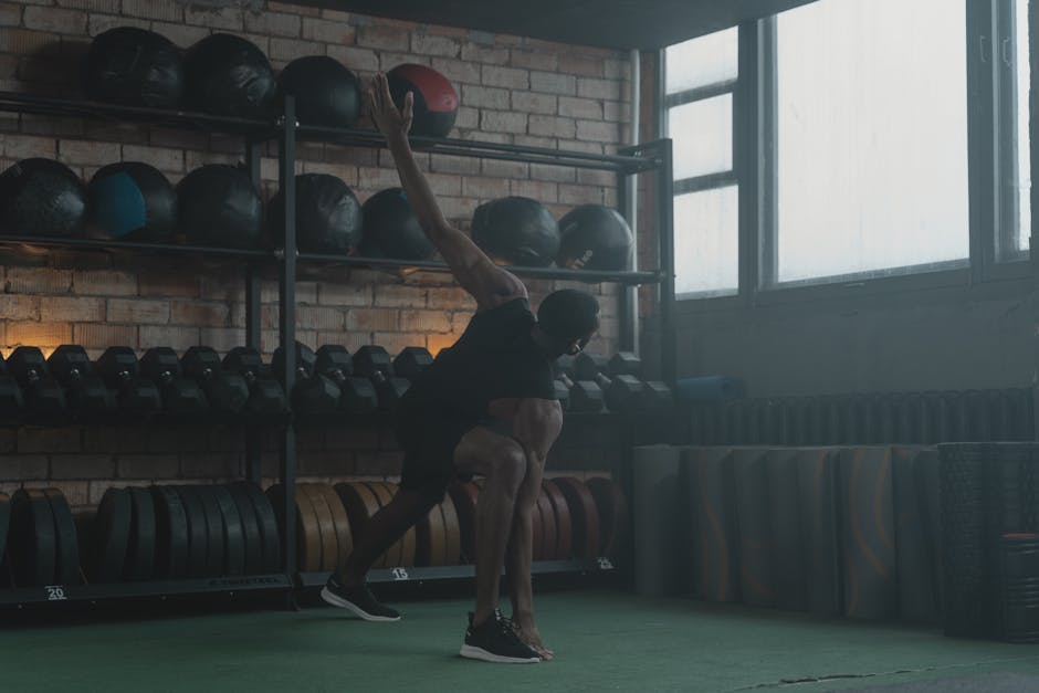 A man performing dynamic stretching exercises in a gym setting with fitness equipment in the background.