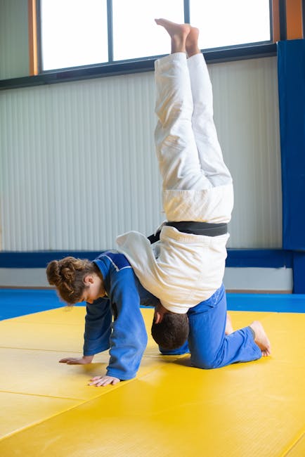 Two martial artists practice judo techniques indoors on a yellow mat.