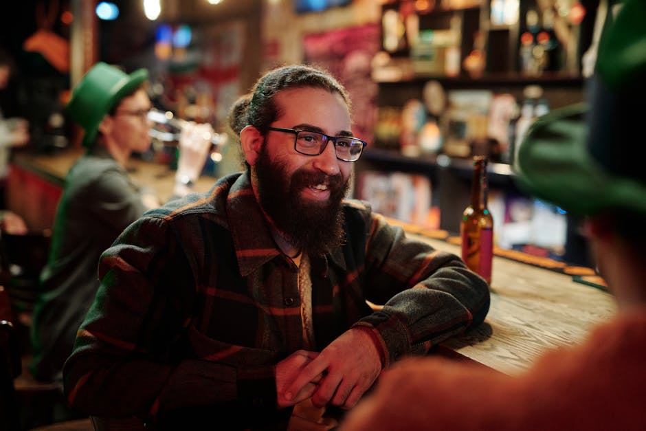 Bearded man in glasses chatting at a rustic bar with friends. Warm indoor ambiance.