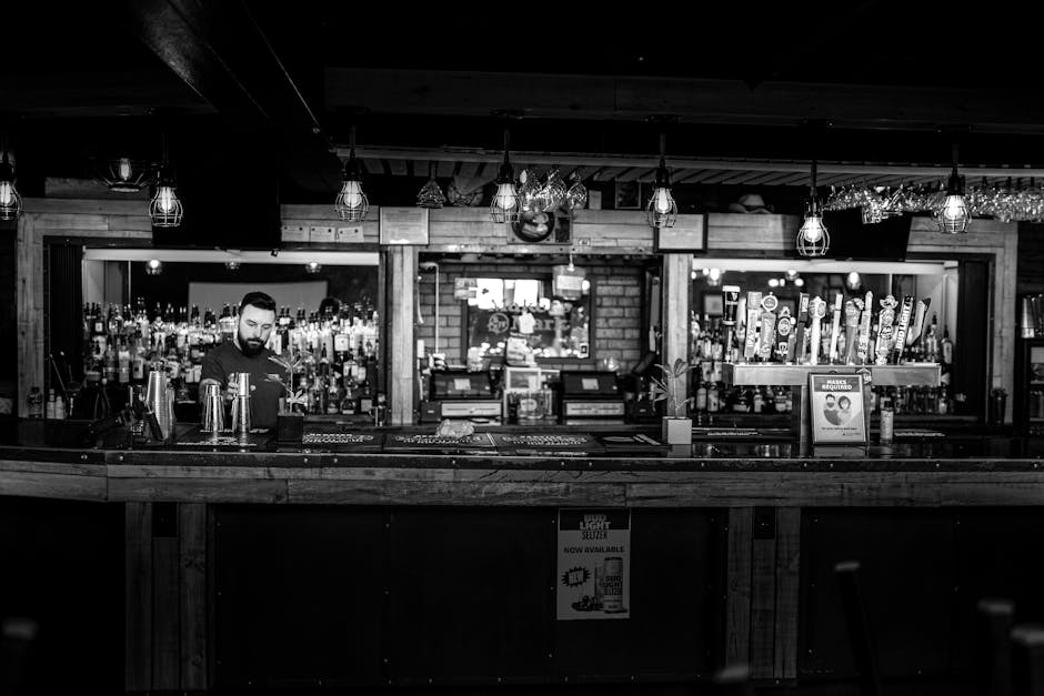A black and white image of a bartender standing at a rustic bar counter in a cozy pub setting.