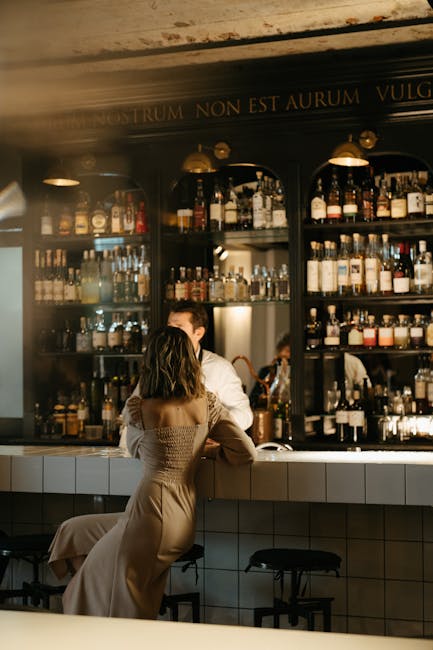 A woman elegantly sits at a modern bar counter filled with bottles, chatting with a bartender.