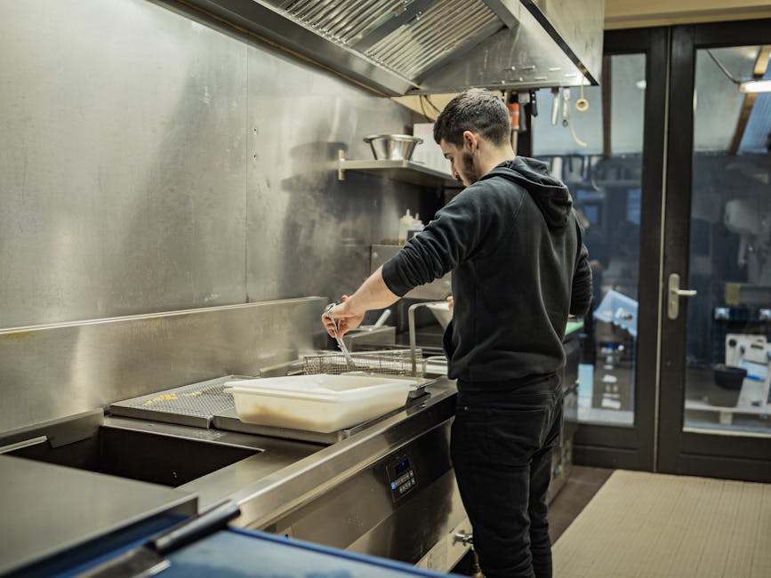 A chef in a black jacket cooks in a modern stainless steel kitchen, frying food indoors.