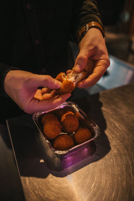 Close-up of hands breaking fried croquettes over a metal table indoors.