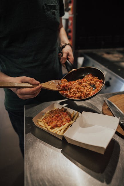 Close-up of a person adding sauce to fries in a takeout box with a frying pan in an indoor kitchen.