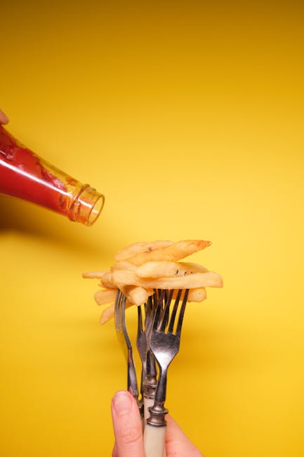 Close-up of fries on forks with ketchup pour against a vibrant yellow background.