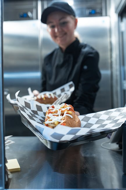 A chef holding a gourmet hot dog with toppings in a professional kitchen setting.