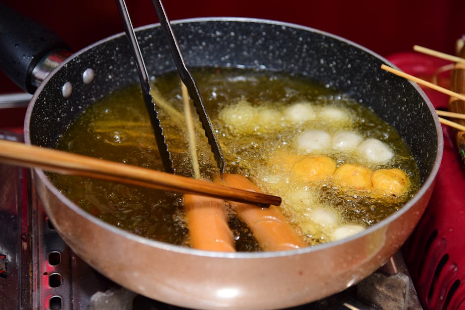 Close-up of a pan frying skewered sausages and fish balls in hot oil.