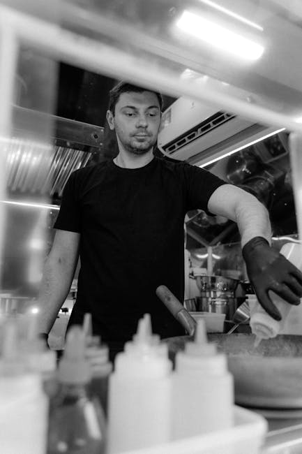 Monochrome image of a chef working in a kitchen with squeeze bottles and stovetop equipment.