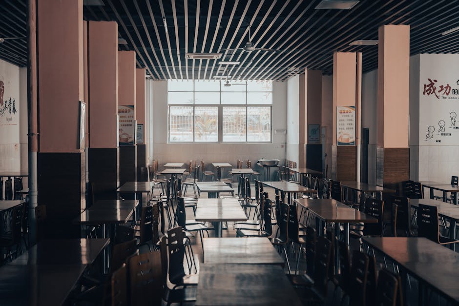 An empty cafeteria featuring wooden tables and chairs, lit by natural light through large windows.