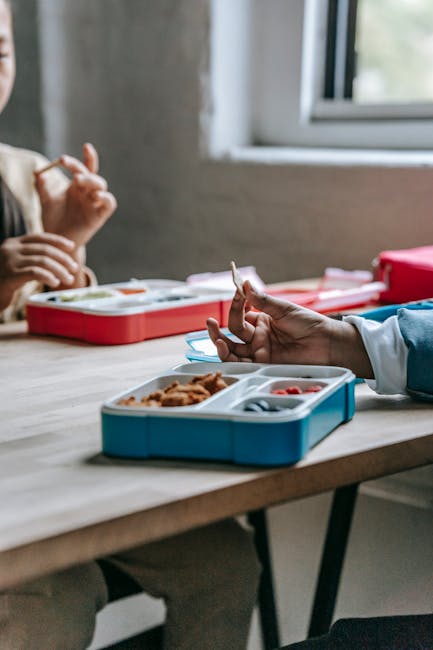 Crop anonymous classmates sitting at wooden table with lunch boxes and eating homemade food