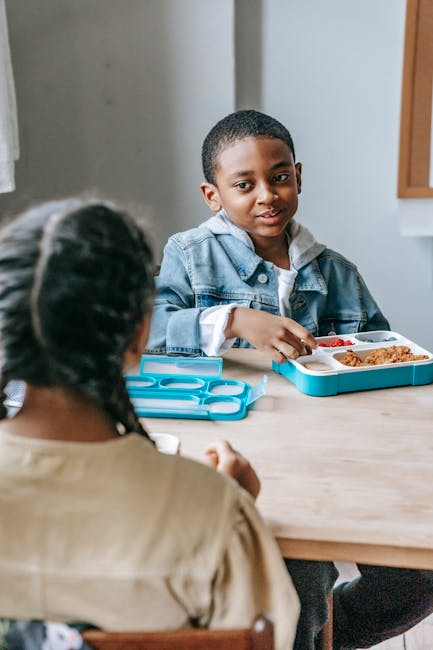 African American schoolchild with school box interacting with crop anonymous ethnic friend at table in classroom