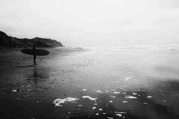 Een surfer staat op een zwart strand met een surfplank, uitkijkend over de zee.