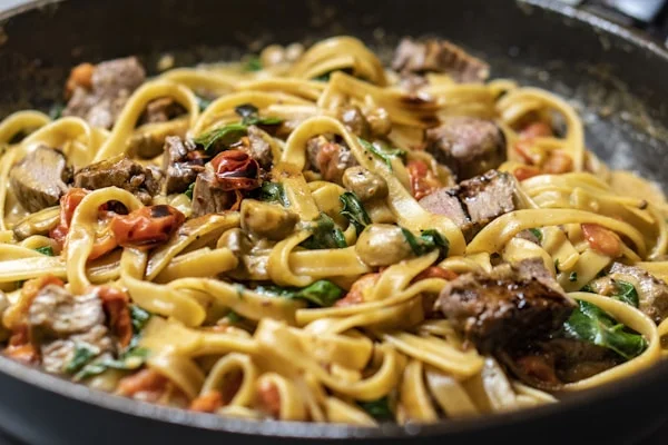 Een smakelijke pasta met stukjes vlees, champignons en tomaat, geserveerd in een zwarte pan.