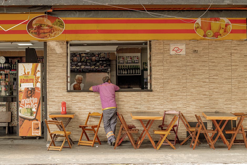 A person in a pink shirt stands outside a street cafe with wooden chairs and tables.