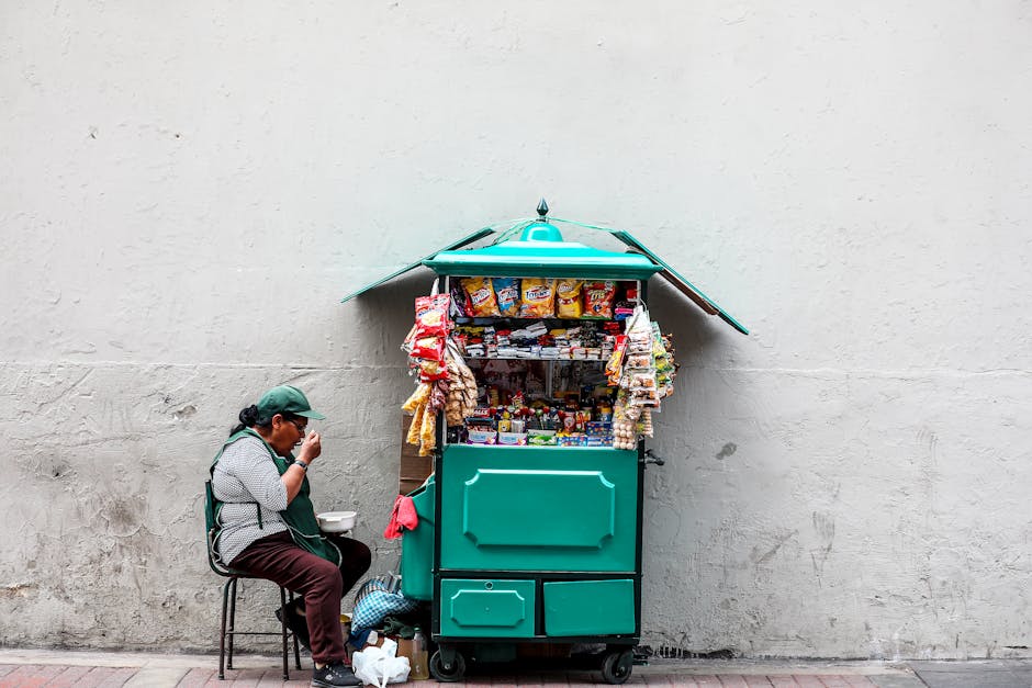 A woman enjoying a meal while sitting next to her snack cart on a city street.
