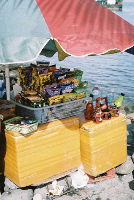 Street vendor stall displaying snacks and drinks under a colorful umbrella by the waterfront.