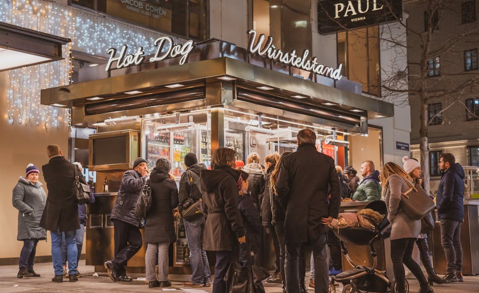 Group of people gathered outside an illuminated Viennese hot dog stall at night in winter.