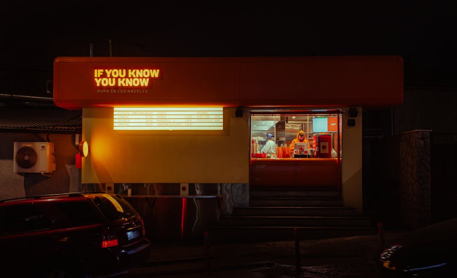 Neon-lit fast food drive-thru at night with visible workers inside.