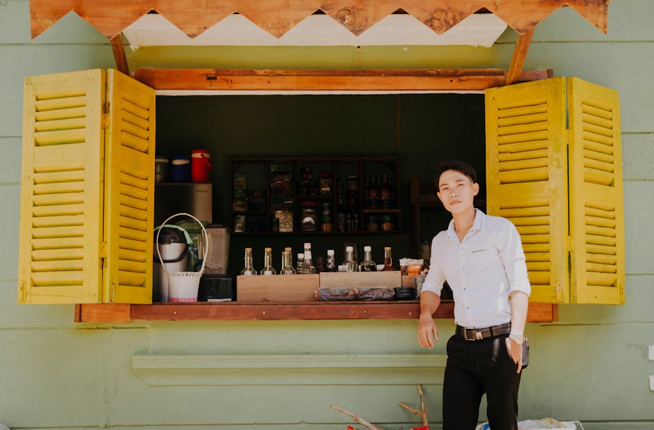 Young dreamy ethnic guy in stylish clothes standing with hand in pocket near window of old building with refreshing drinks for sale while looking at camera