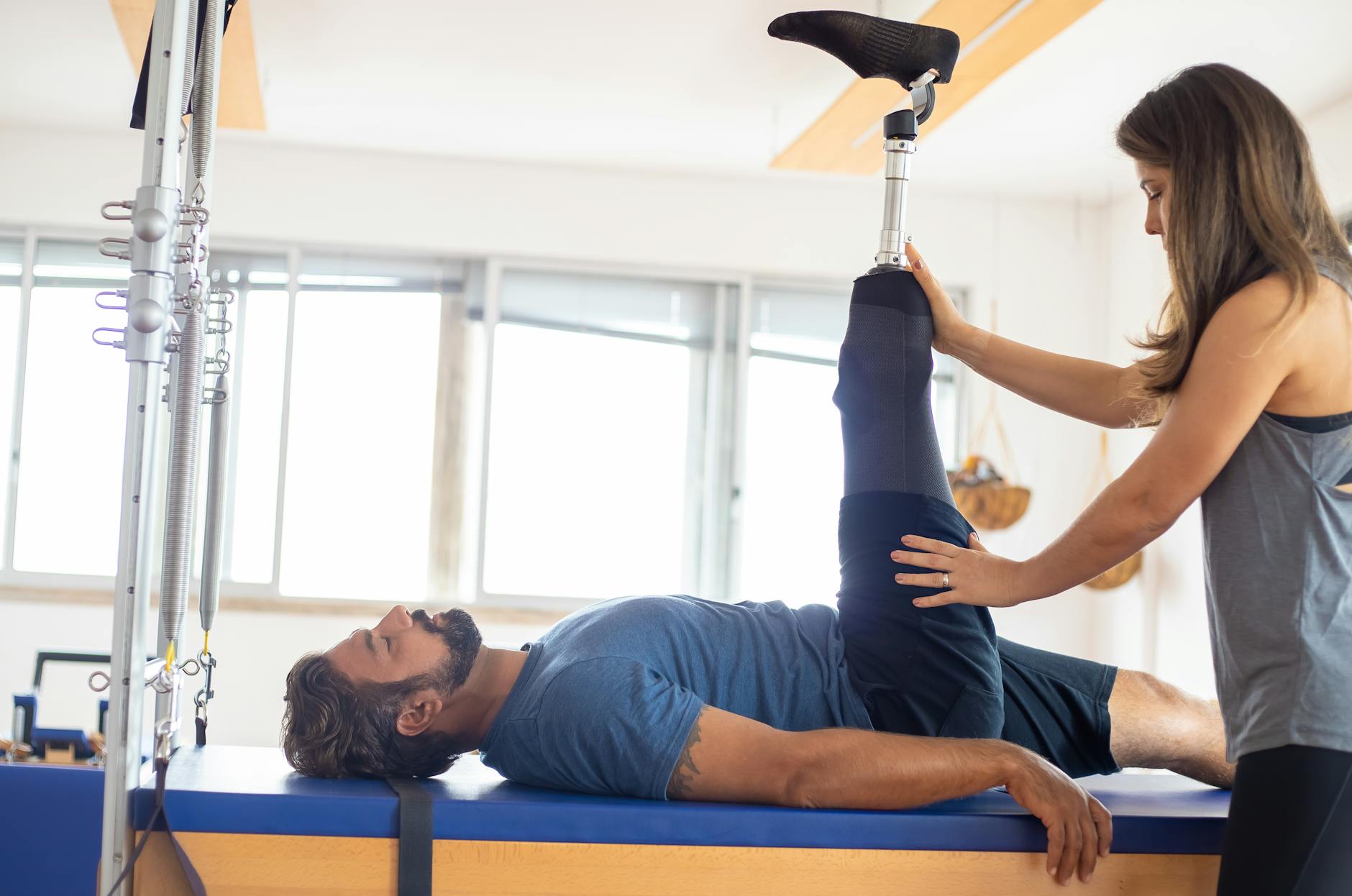 Man met beenprothese krijgt fysiotherapie op een pilates apparaat, geholpen door een vrouw.